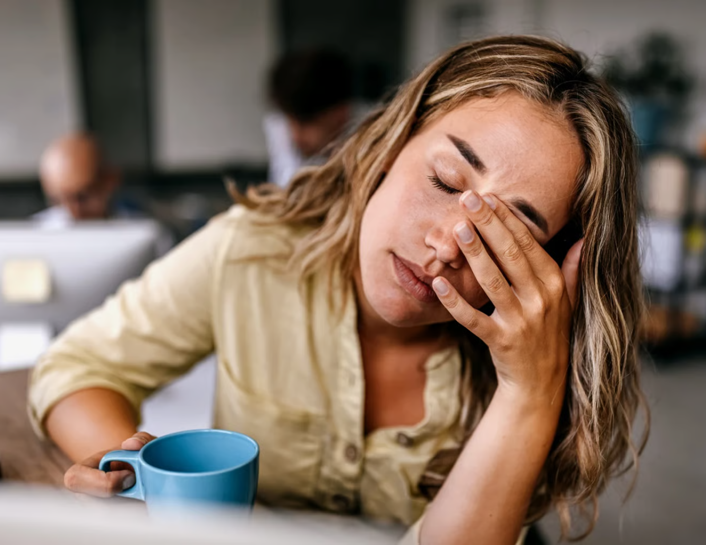 Tired woman holding a blue mug, sitting at a desk.
