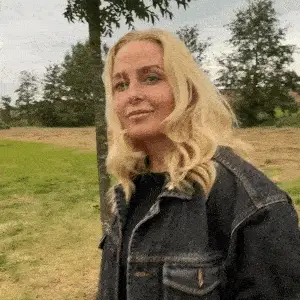 A blonde woman in a denim jacket smiles and turns her head slightly while standing outdoors.