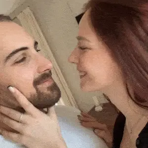 A close-up of a smiling woman with long red hair kissing a bearded man on the nose.