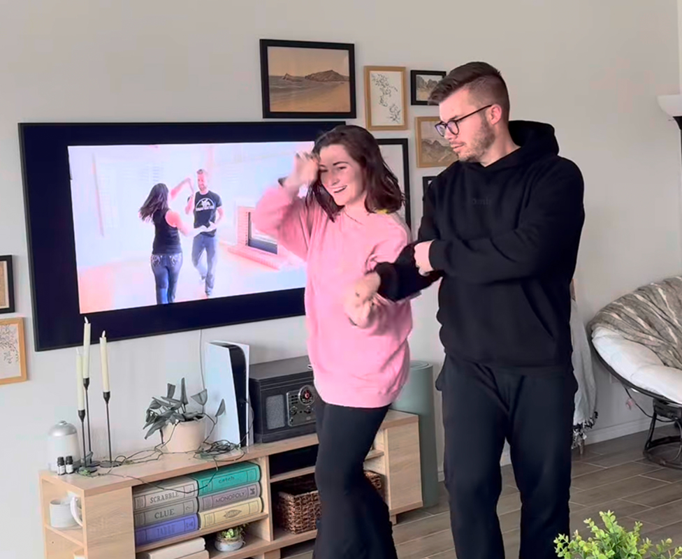 A couple dancing in a living room with a TV showing dancers.
