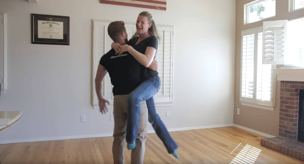A man and woman dance together joyfully in a cozy living room, surrounded by warm lighting and comfortable furniture.