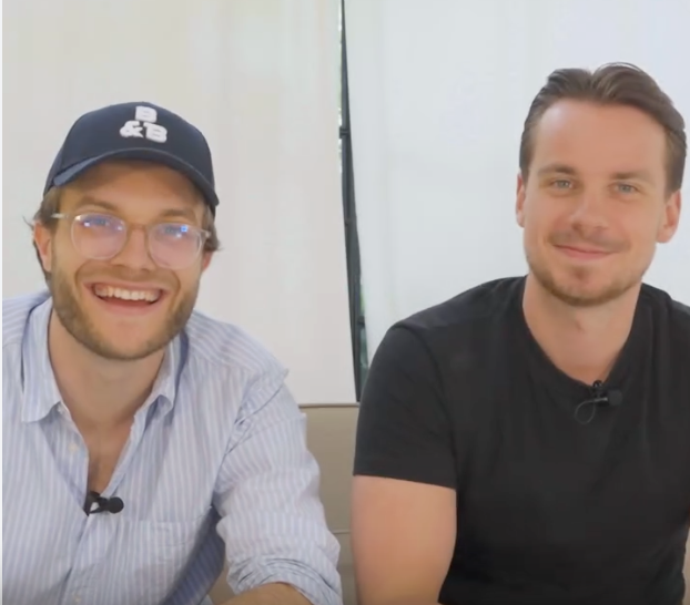Two men sitting and smiling indoors, one wearing a cap.