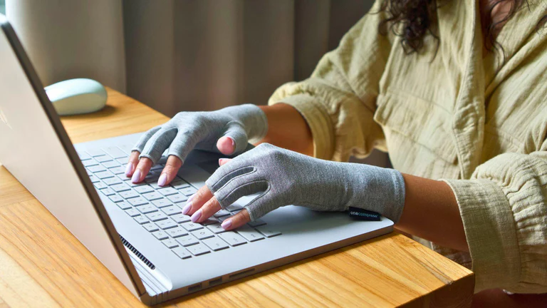 Person wearing gray gloves typing on a laptop at a wooden desk.