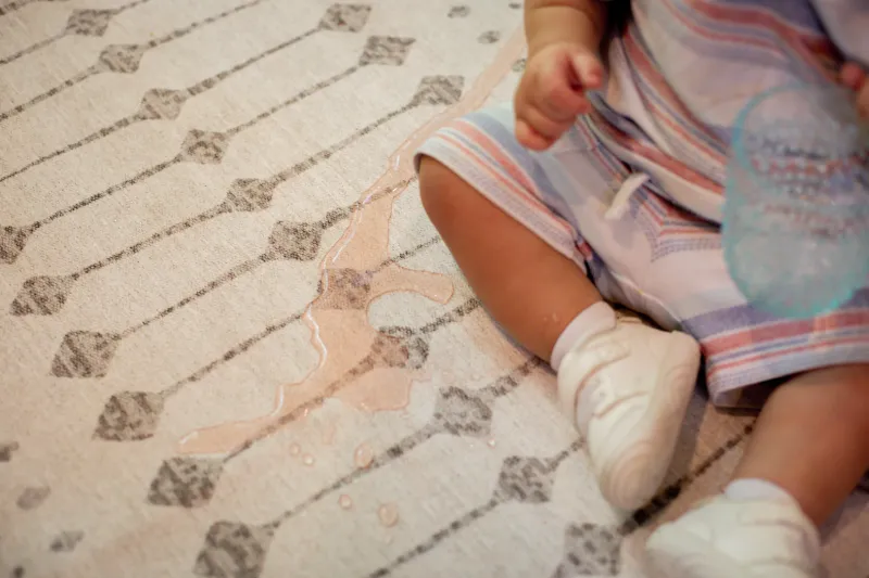 A baby sits on a patterned rug next to a liquid spill.