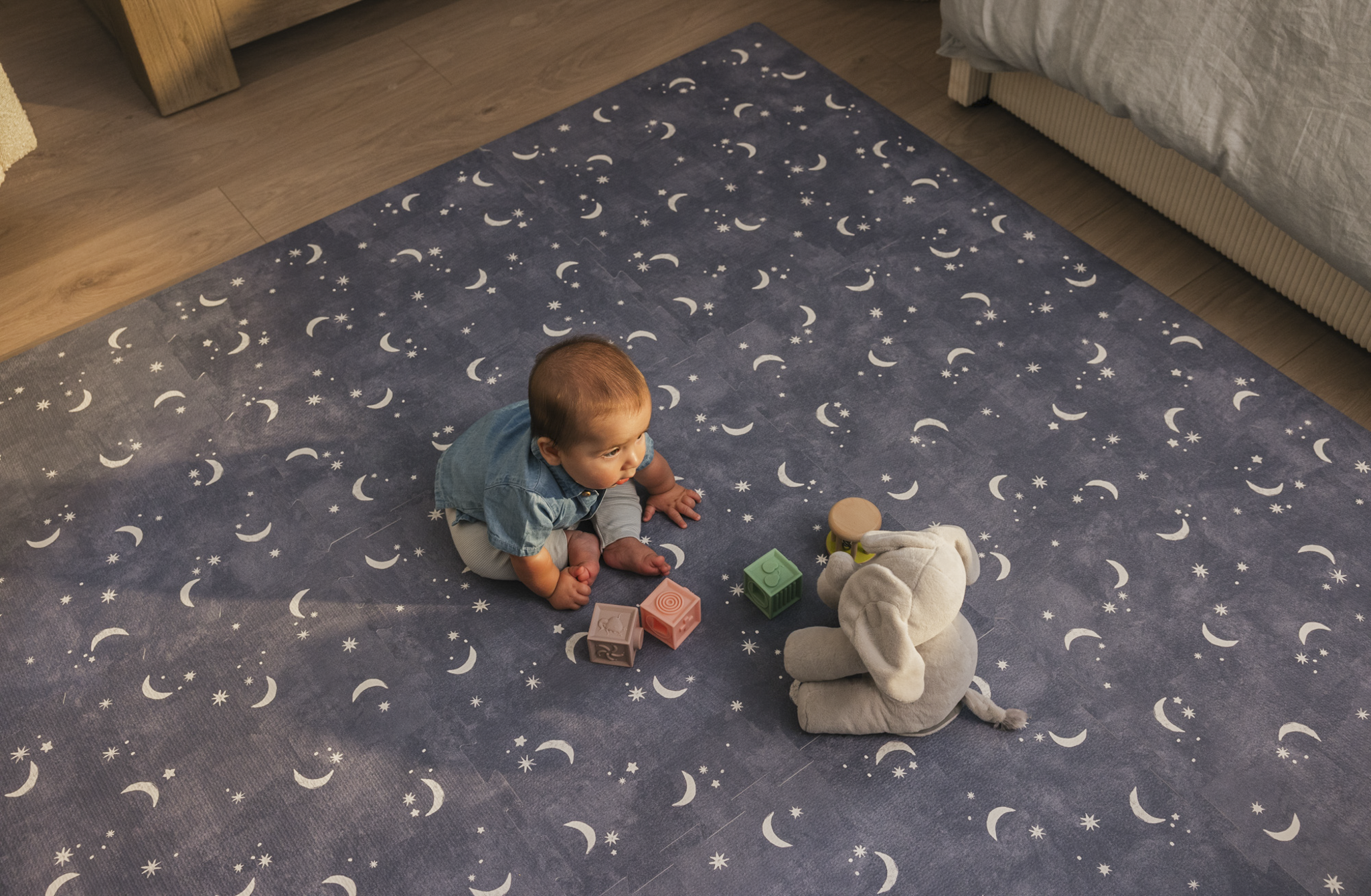 Baby sitting on a moon-patterned mat with toys and a stuffed elephant.