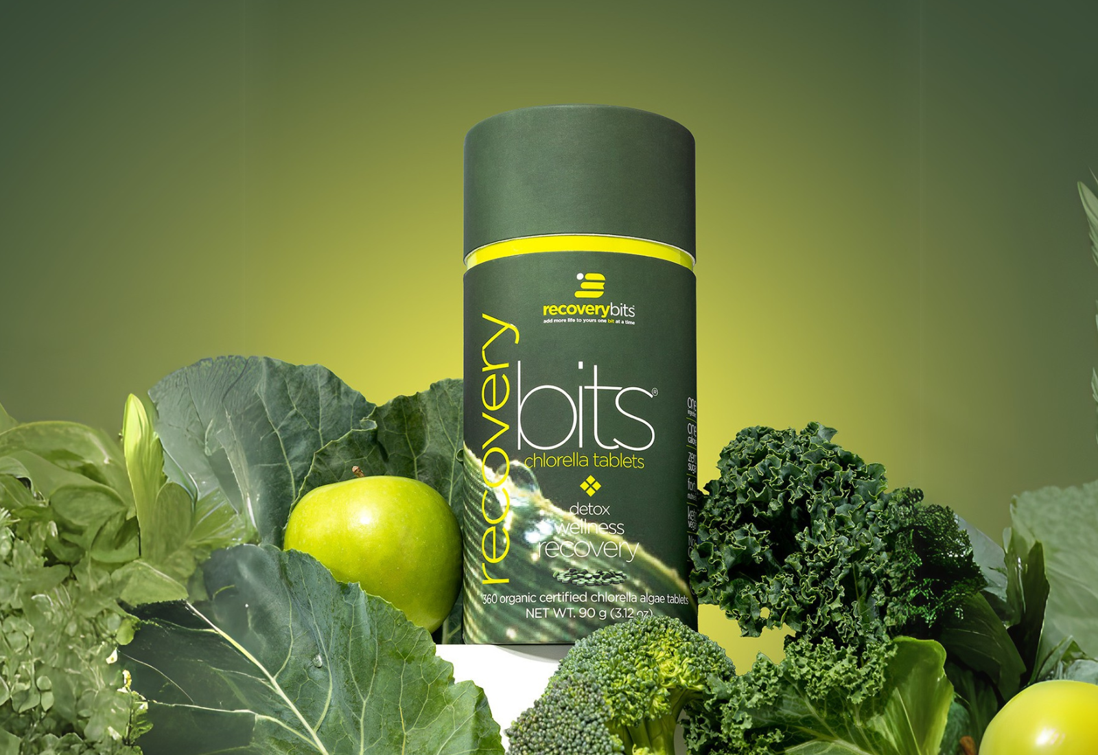 Container of chlorella tablets surrounded by green vegetables and an apple.