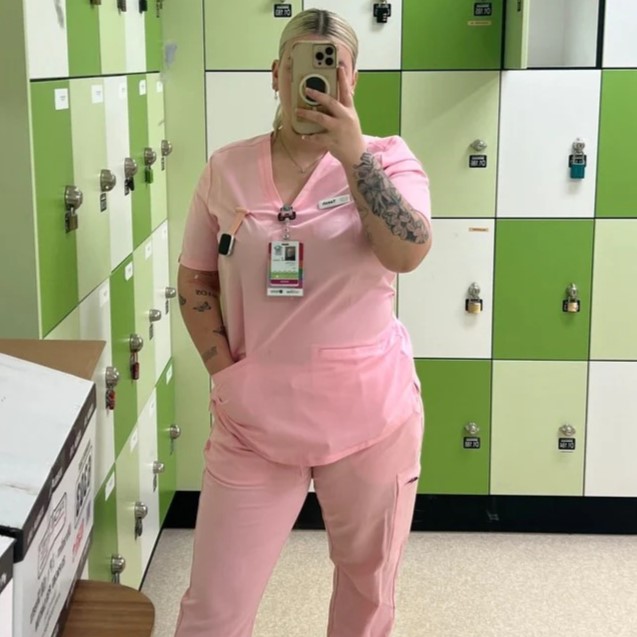 Person in pink scrubs taking a mirror selfie in a locker room.