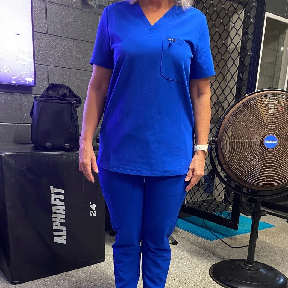 Person in blue scrubs standing in a gym with a fan and equipment.