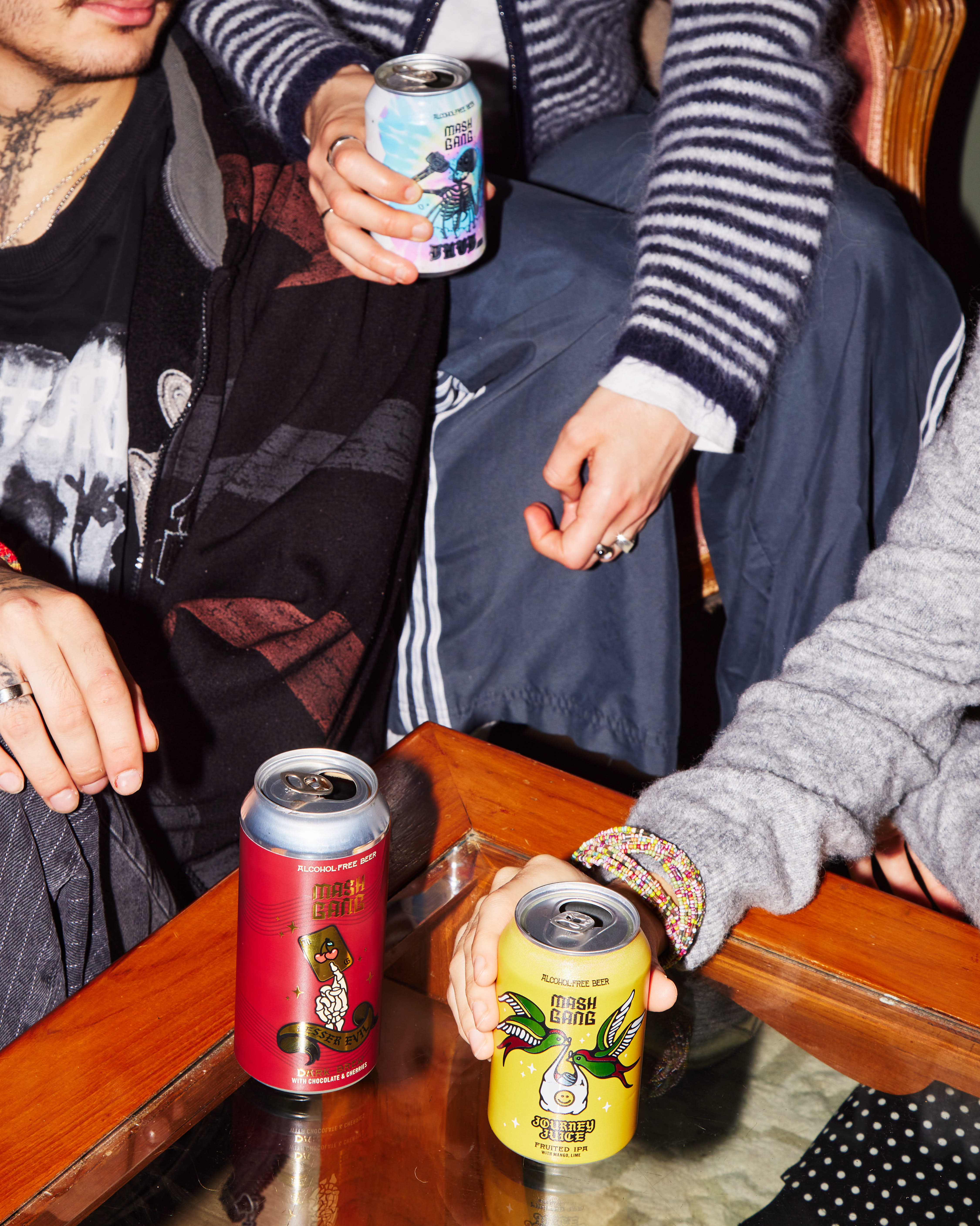 People holding colorful Mash Gang beverage cans around a wooden table.