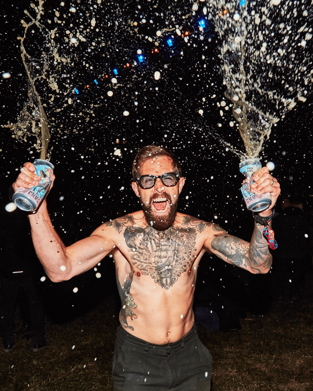 Shirtless person with tattoos spraying two cans of liquid in the air at night.