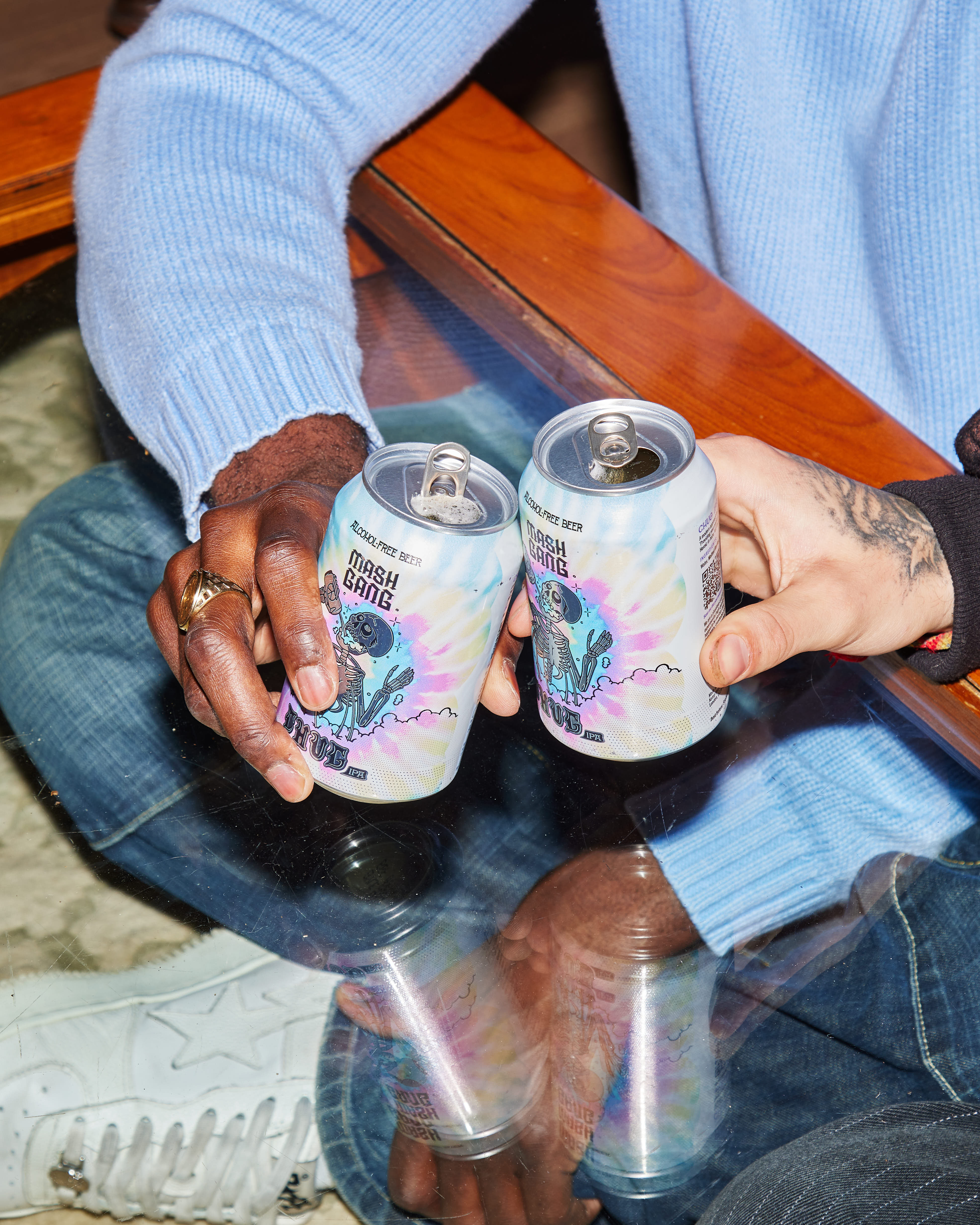 Two hands holding and clinking cans of Mash Gang alcohol-free beer over a glass table.