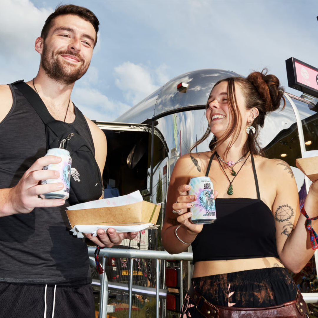 Two people smiling, holding food and drinks in front of a shiny trailer.