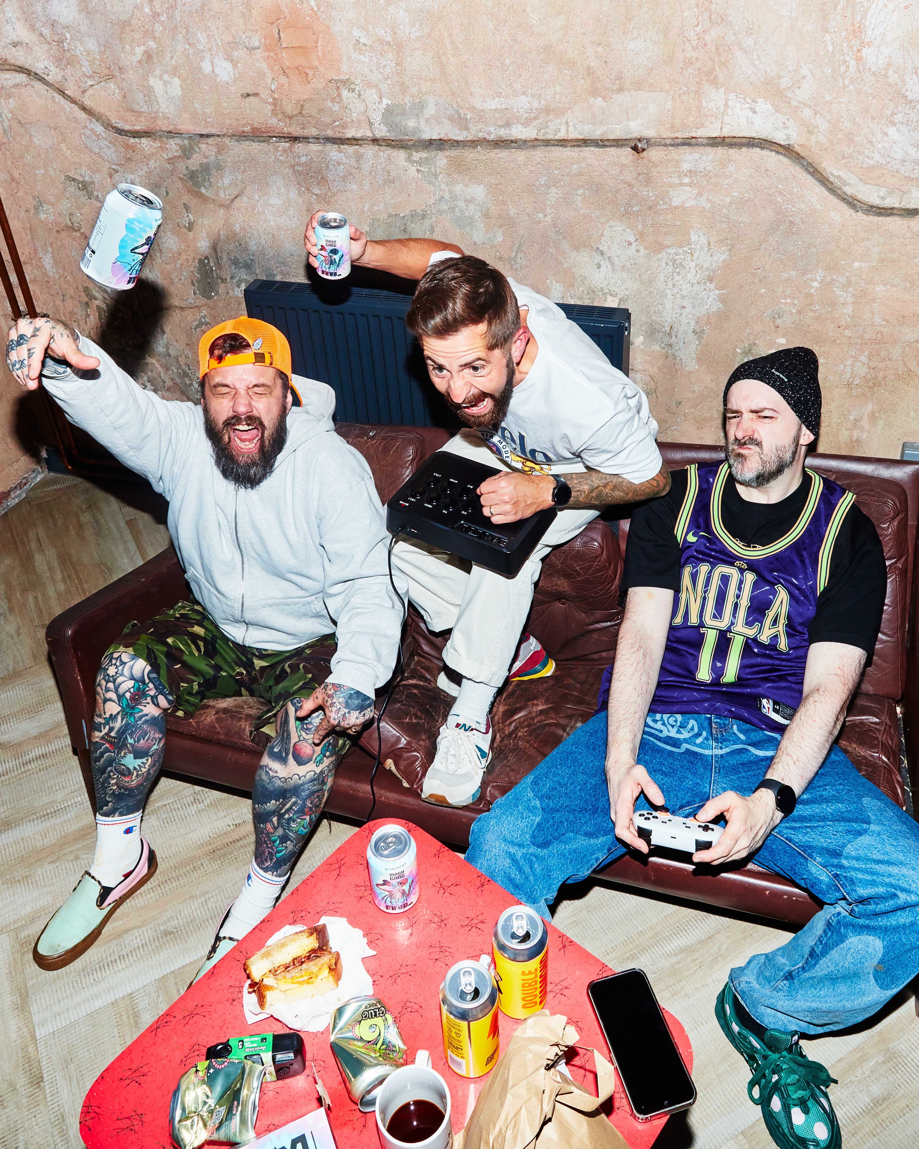 Three men on a couch, holding drinks and gaming controllers, with snacks on a table.