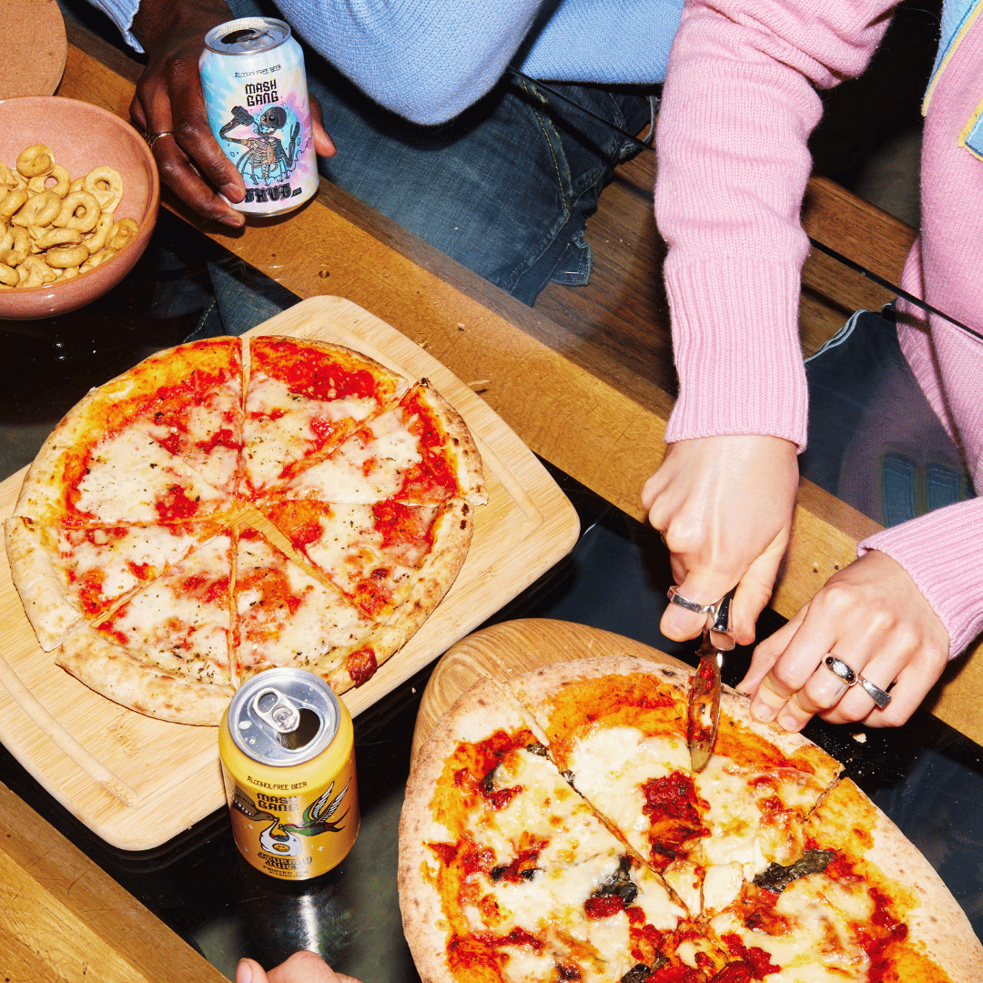 Two pizzas on wooden boards, person slicing, drinks and snacks on table.