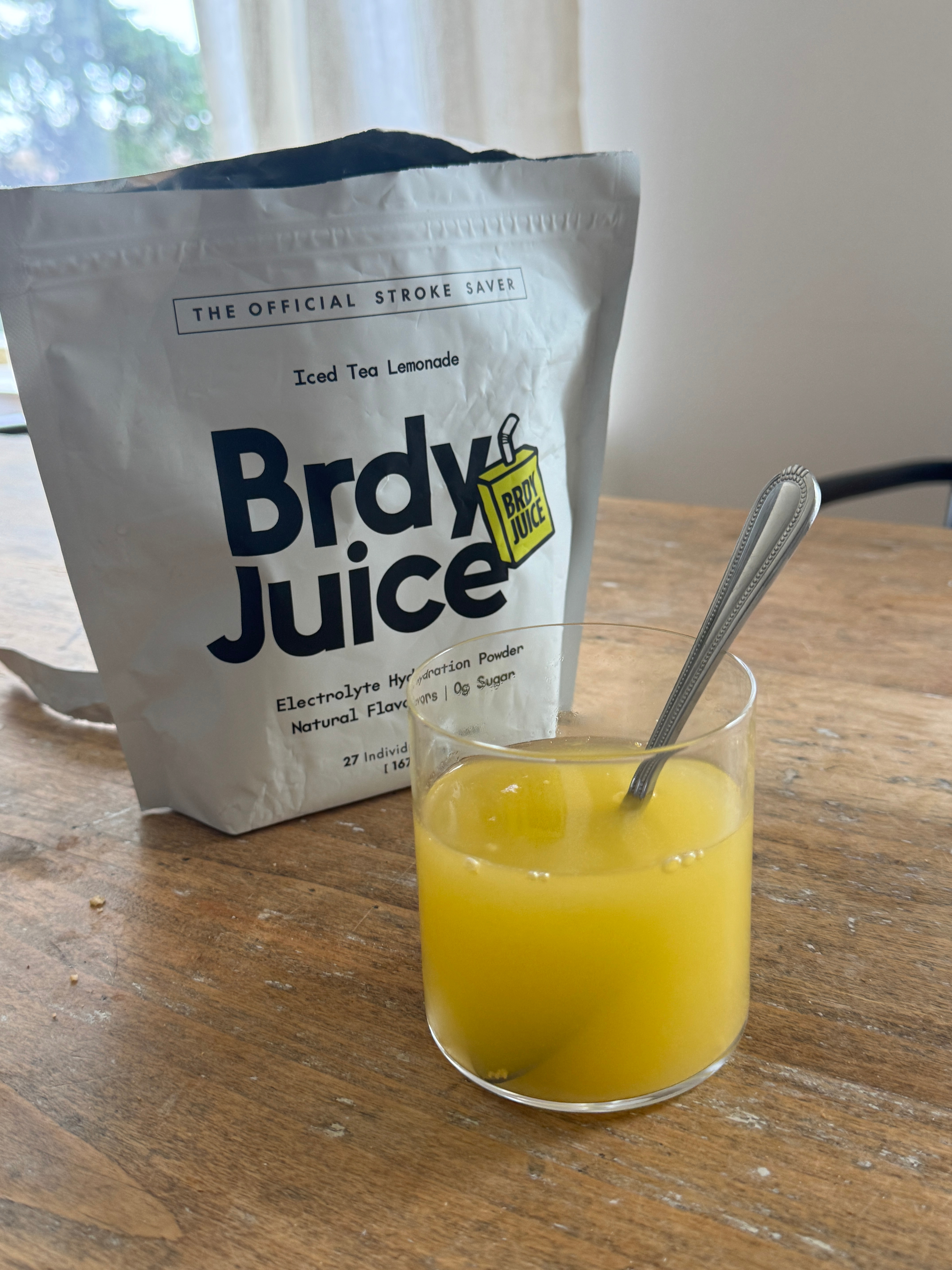 Packet of electrolyte powder and a glass of yellow drink on a wooden table.