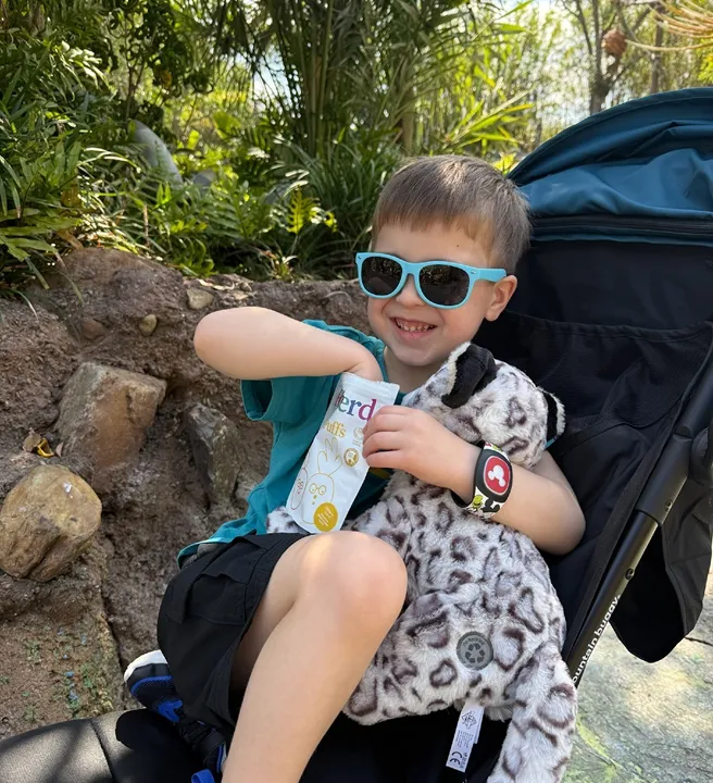Child in sunglasses sits in a stroller, smiling with a stuffed animal and snack.