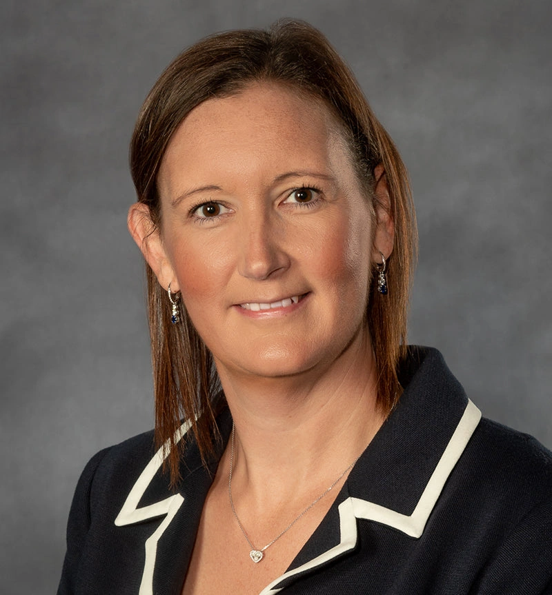 Person with short brown hair in a dark suit, smiling against a gray background.