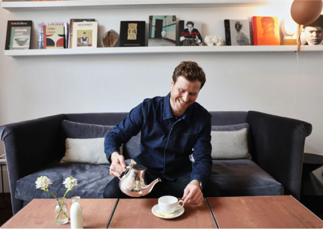 Man pouring tea in a cozy living room with books on a shelf.