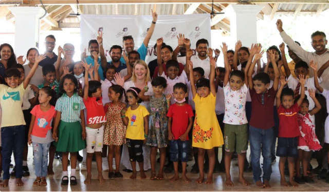 A group of children and adults smiling and raising their hands in celebration indoors.