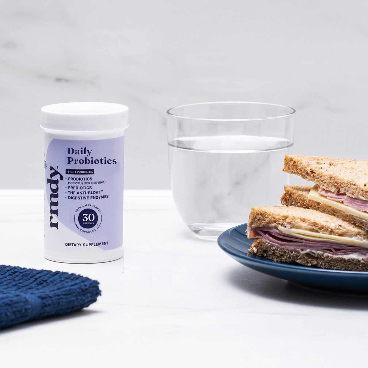Probiotic bottle, glass of water, and sandwich on a blue plate on a white surface.