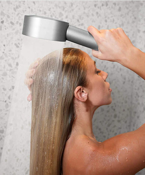 Person washing hair under a showerhead in a tiled bathroom.