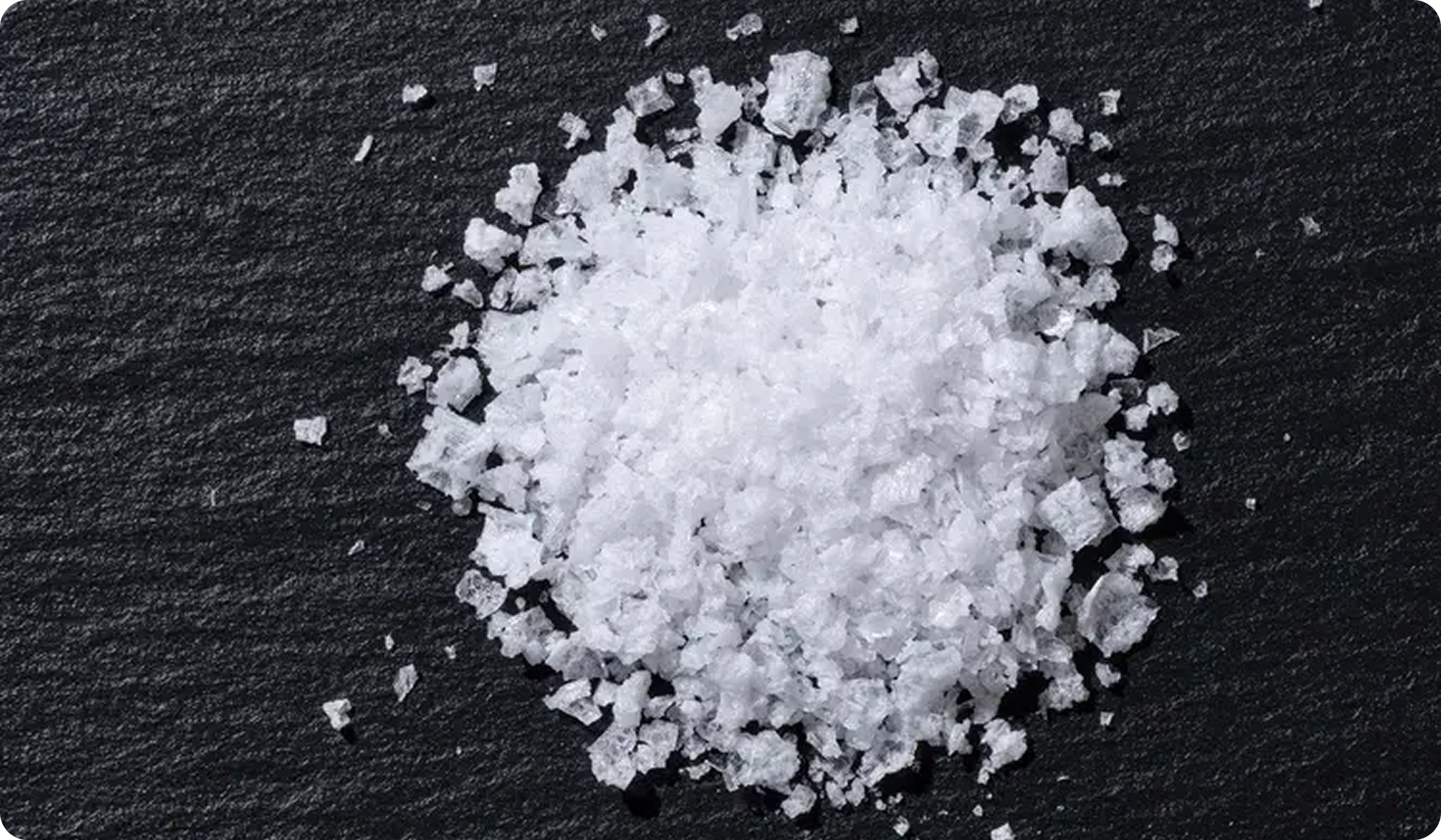 A close-up overhead shot of a pile of coarse white salt on a dark textured surface.