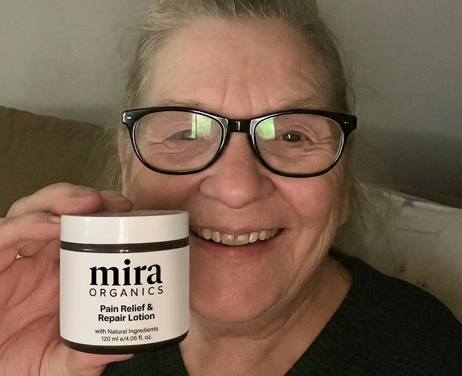 A smiling woman with glasses holds up a white jar of Mira Organics Pain Relief & Repair Lotion.