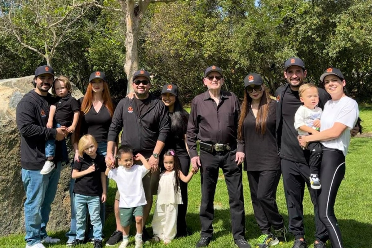 A large multi-generational family posing for a group photo outdoors in a park, many wearing matching hats.
