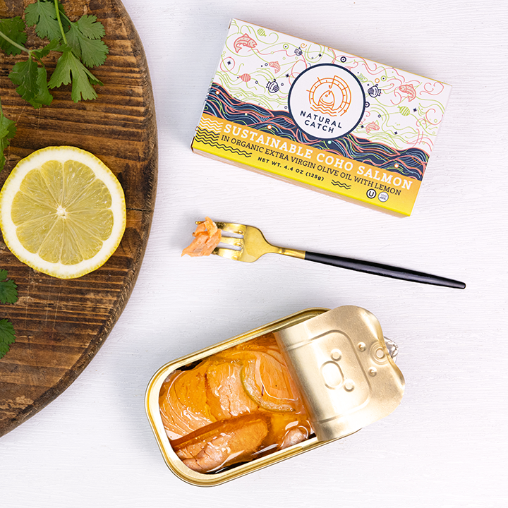 An open tin of coho salmon next to its box, a lemon slice, cilantro, and a fork.
