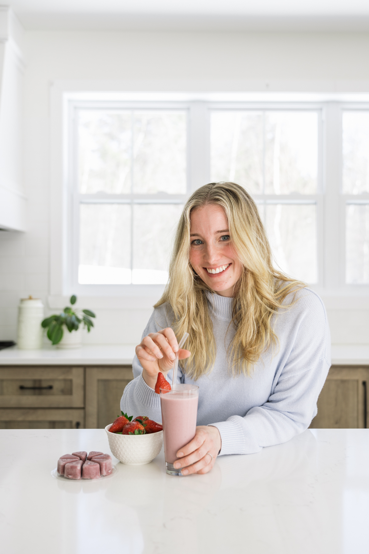 Woman smiling and holding a strawberry smoothie in a bright kitchen.