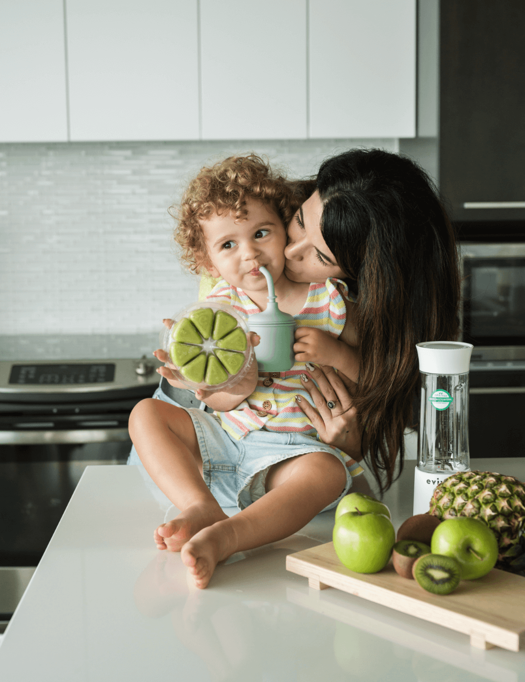 Woman kissing child holding a drink on kitchen counter with fruits.