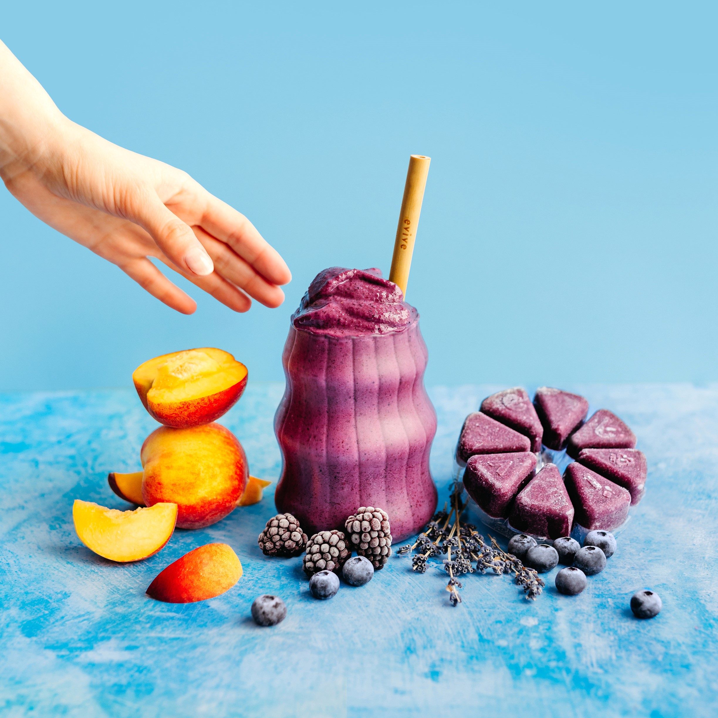 Purple smoothie with a straw, surrounded by fruits and a hand reaching out.