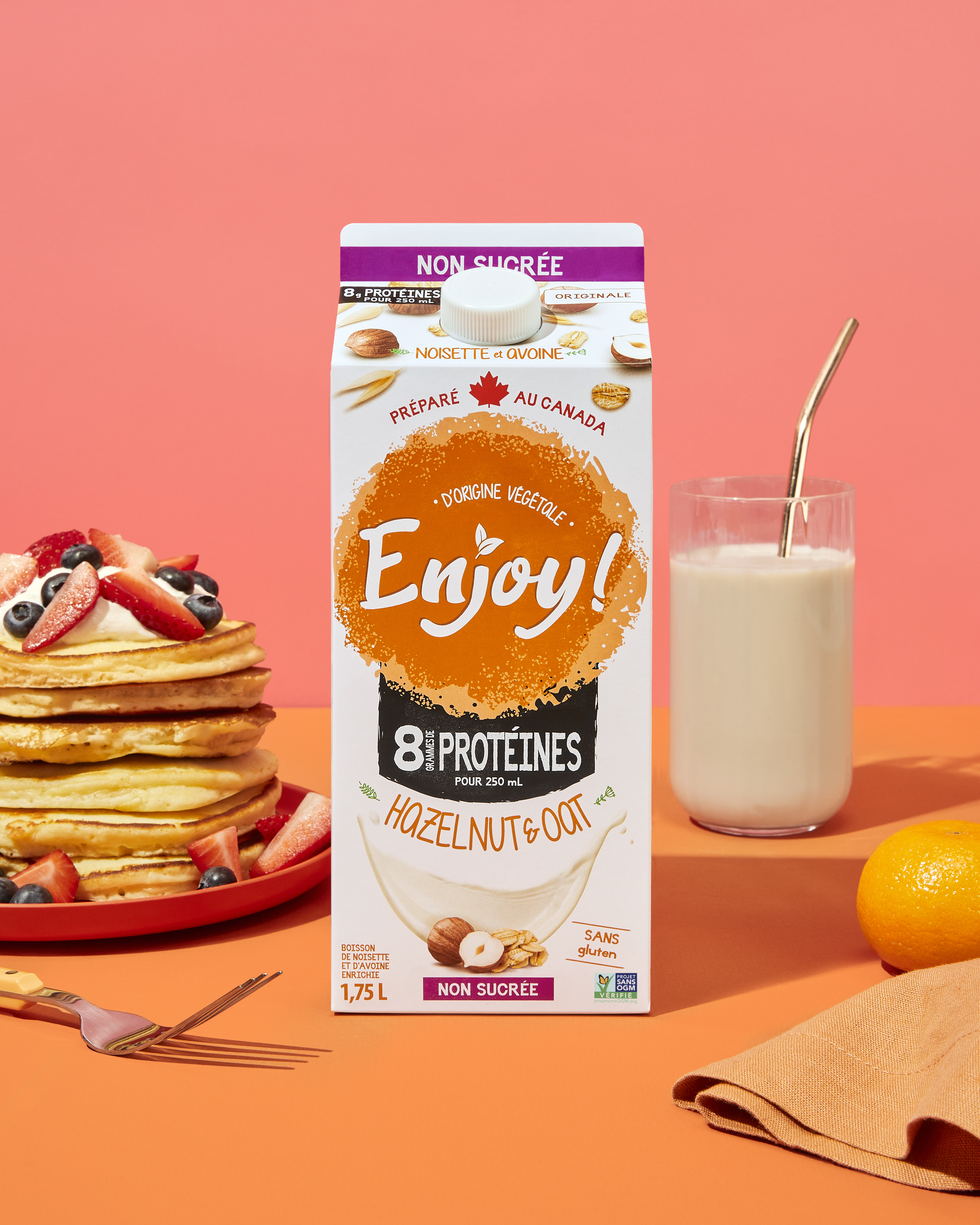 Carton of hazelnut oat milk with pancakes, glass, and orange on a pink background.