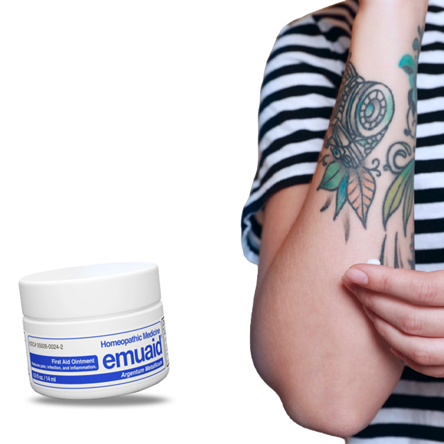 Una persona con un tatuaje en el brazo se aplica crema, junto a un frasco flotante de pomada « Emuaid ».