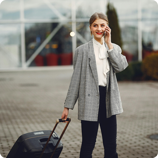 Woman in a plaid blazer talking on phone, pulling a suitcase outdoors.