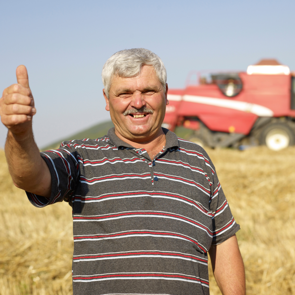 Smiling man in a striped shirt giving a thumbs-up in a field with a red harvester.