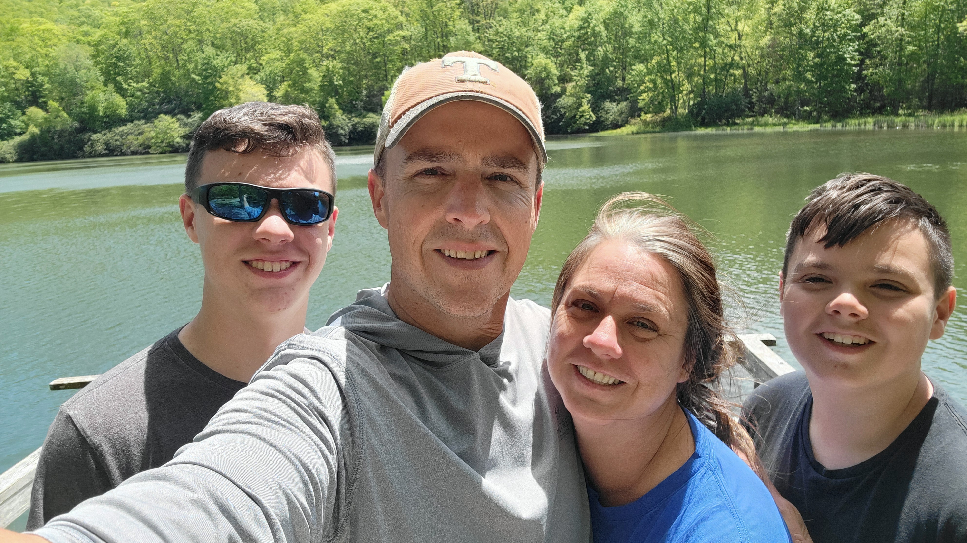 Family of four smiling by a lake with trees in the background.