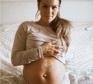 A pregnant woman sits on a bed with a heart drawn in lotion on her bare belly.