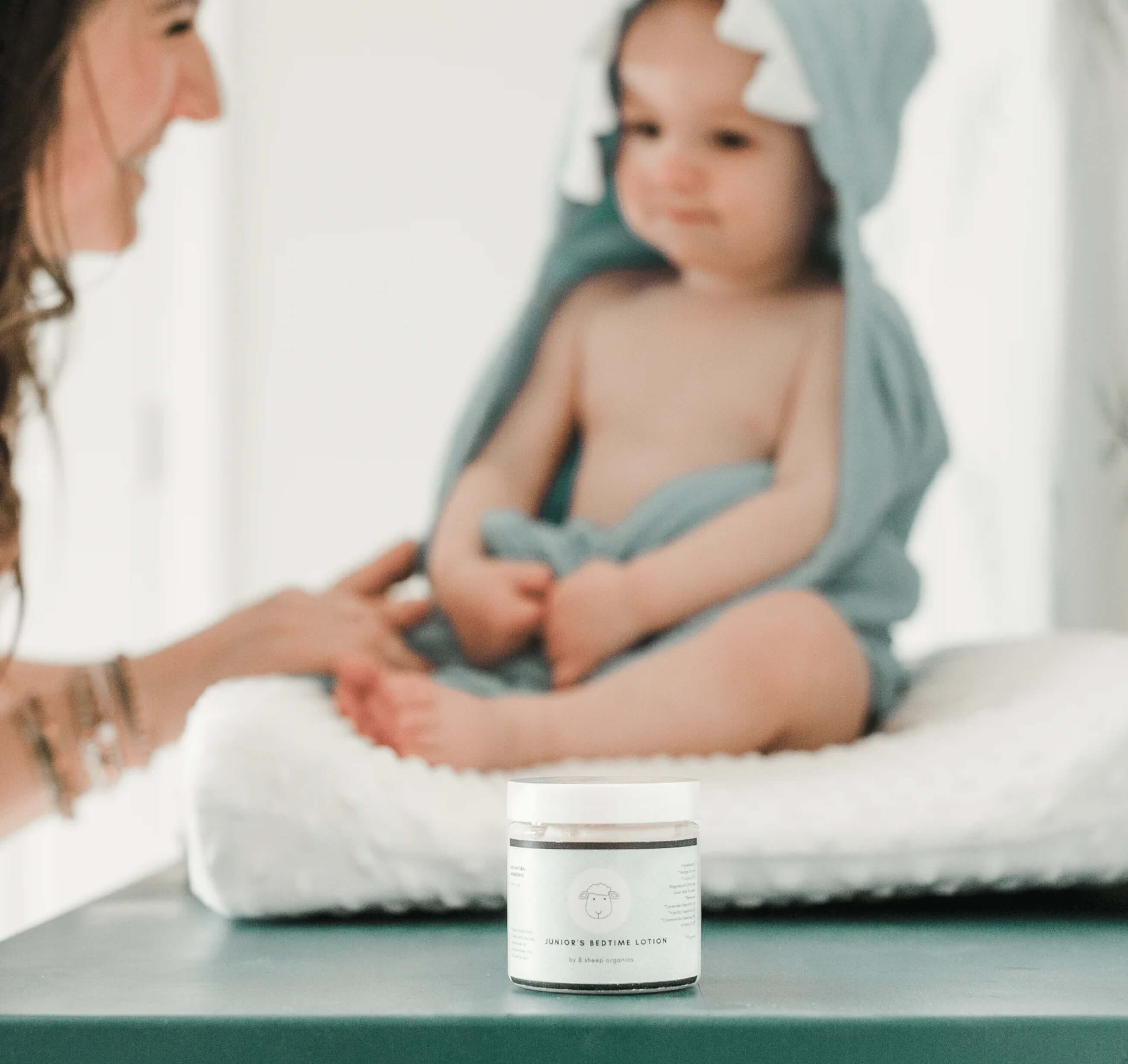 A jar of lotion in focus with a baby in a hooded towel in the background.