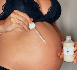 A pregnant person in a lace bra applies a liquid from a dropper to their bare belly.