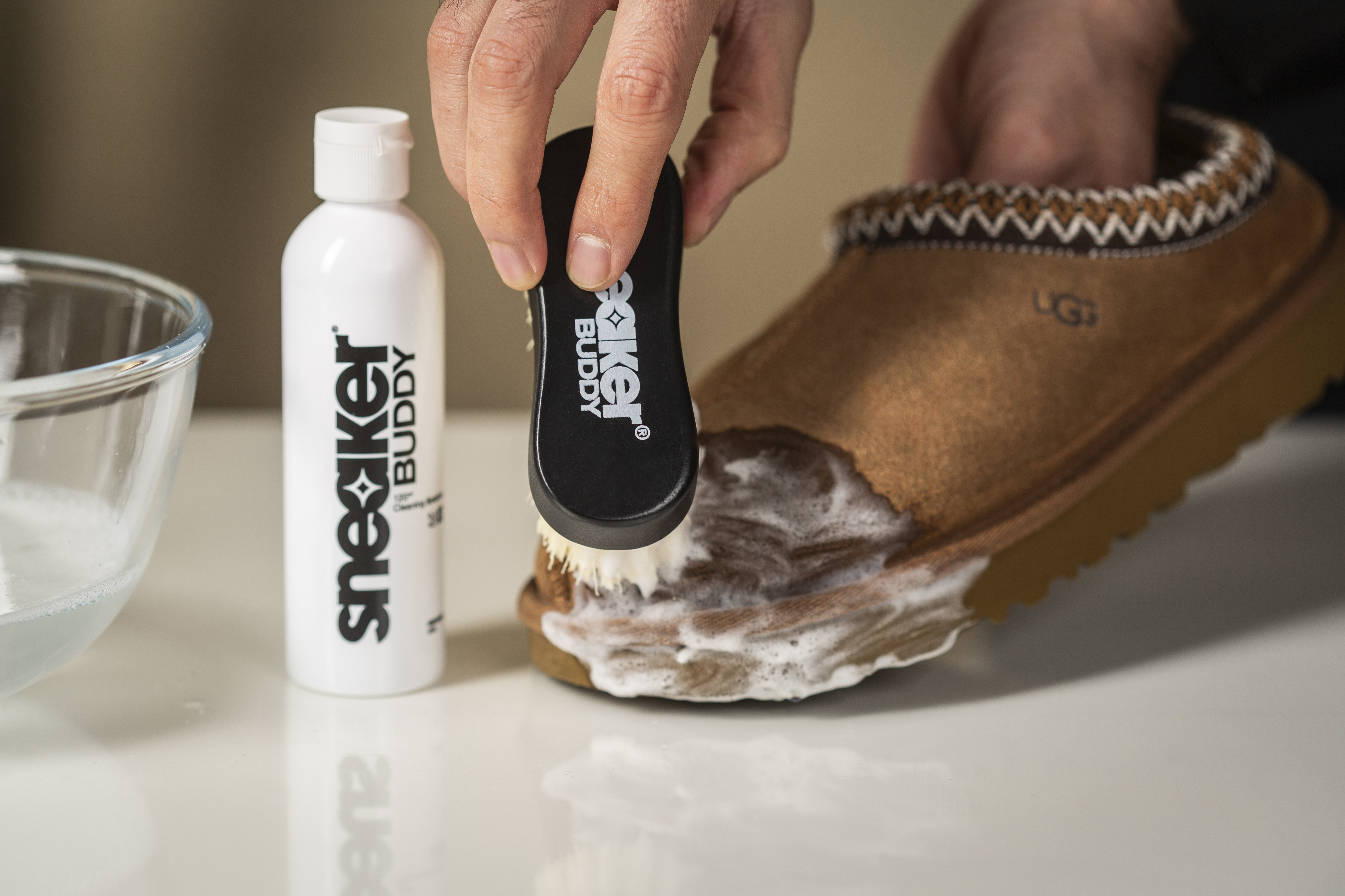A person cleans a brown suede UGG slipper with a Sneaker Buddy brush and cleaning foam.