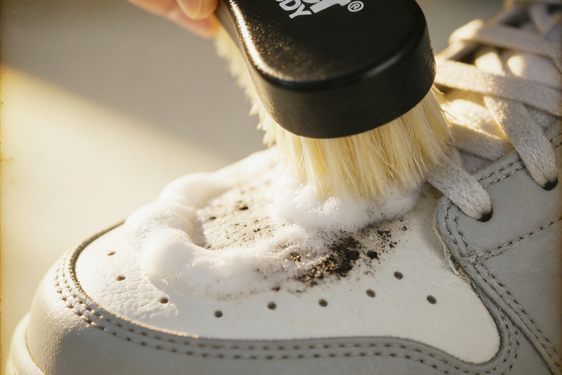 A close-up shot of a person scrubbing a dirty white sneaker with a brush and cleaning foam.