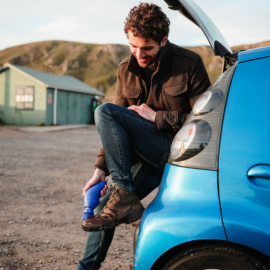 Person cleaning muddy boot with a brush near a blue car.