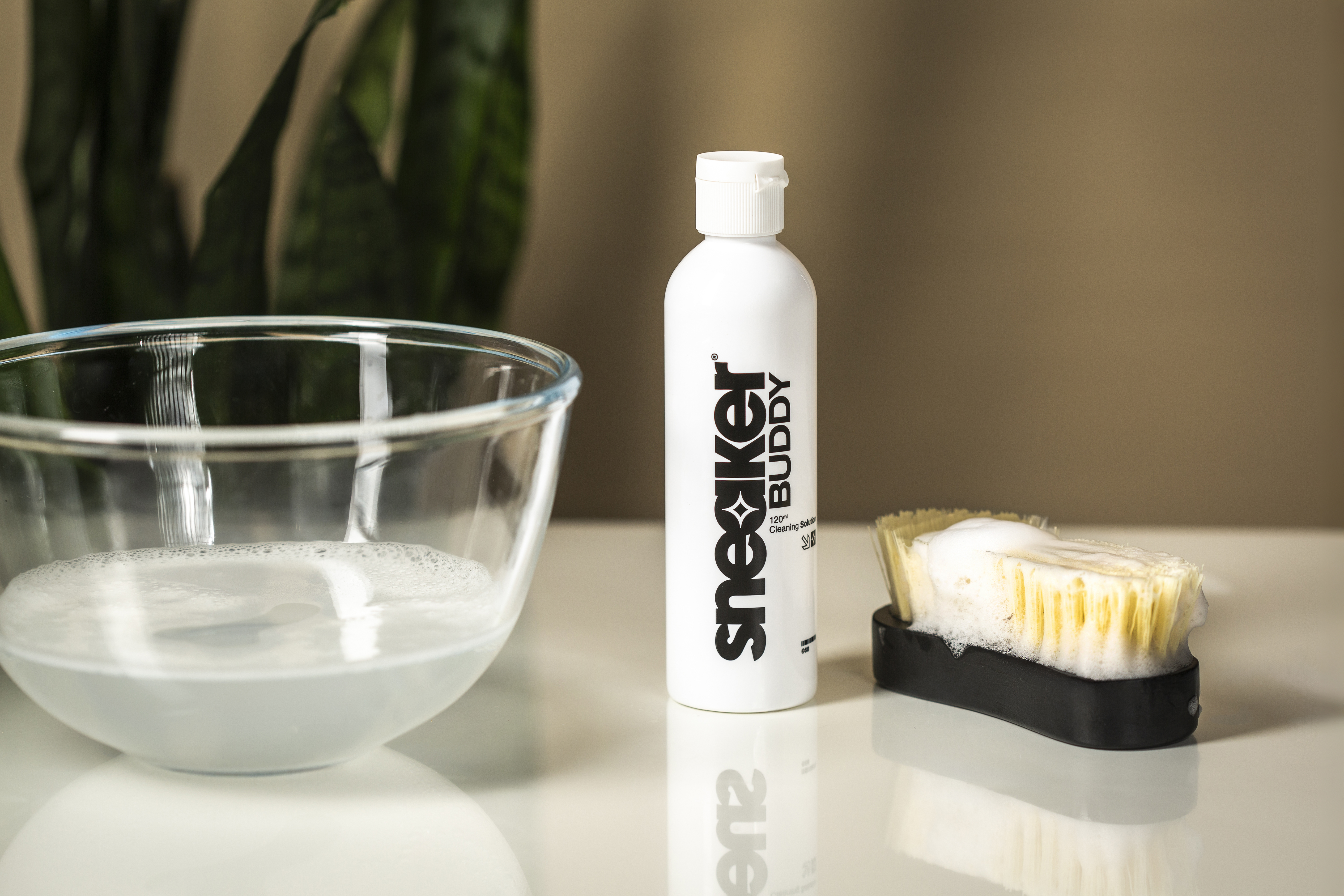A bottle of Sneaker Buddy cleaning solution next to a sudsy brush and a bowl of soapy water.