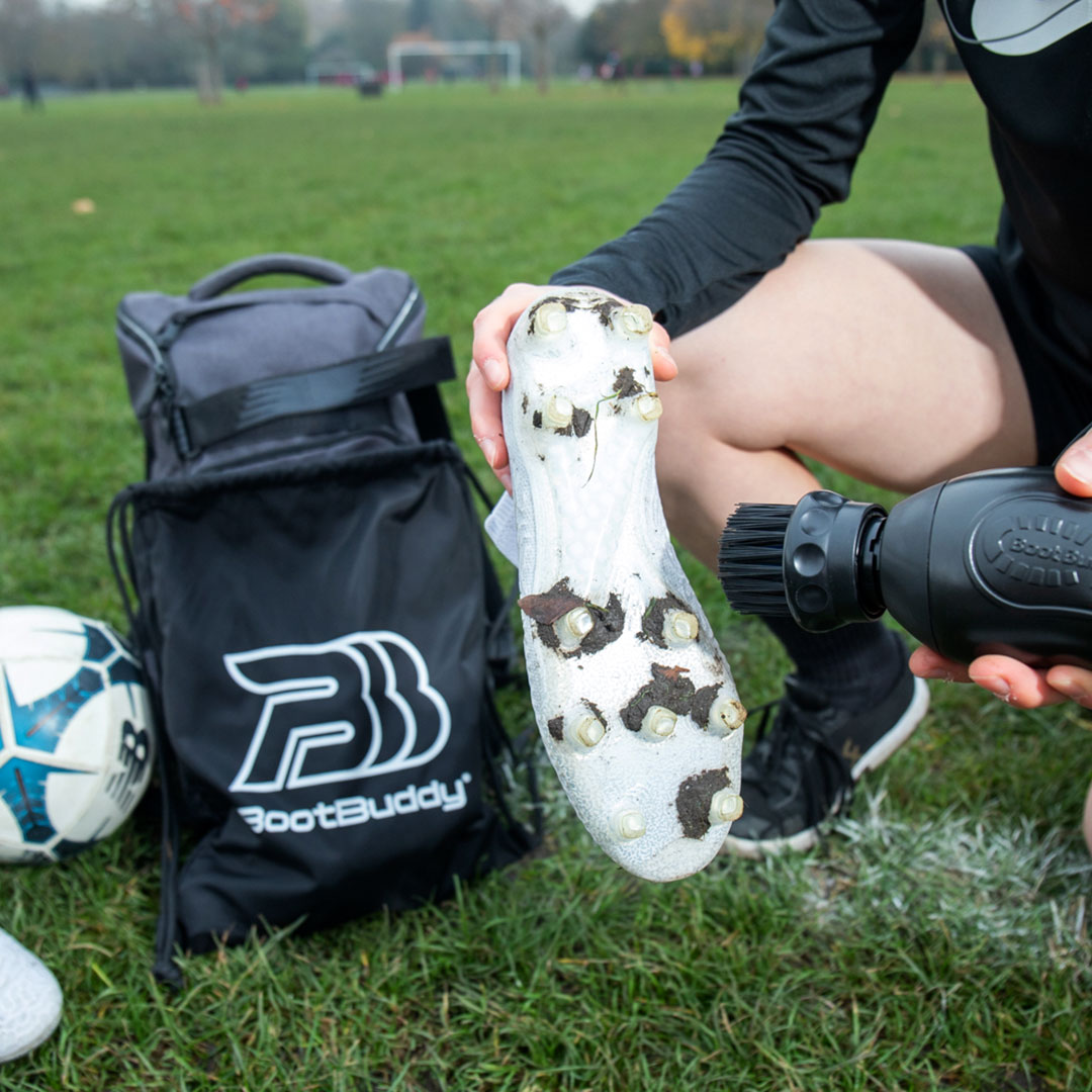 Person cleaning soccer cleats with a tool next to a soccer bag and ball on grass.
