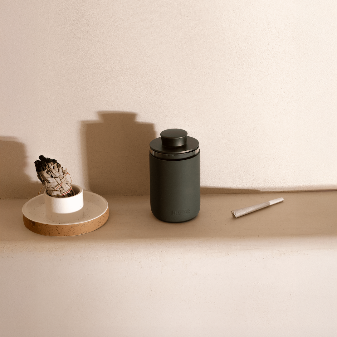 A dark green canister, a bundle of sage in a holder, and a joint on a shelf.