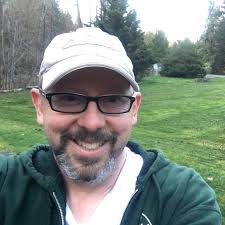 A man wearing glasses and a cap, smiling outdoors.