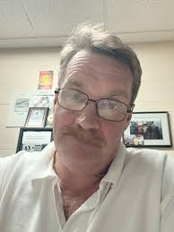 A man with a mustache and glasses, wearing a white polo shirt, takes a selfie in an office.