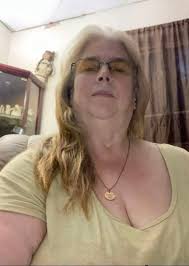 A close-up photo of a woman with blonde hair and glasses, wearing a necklace and a light-colored shirt.