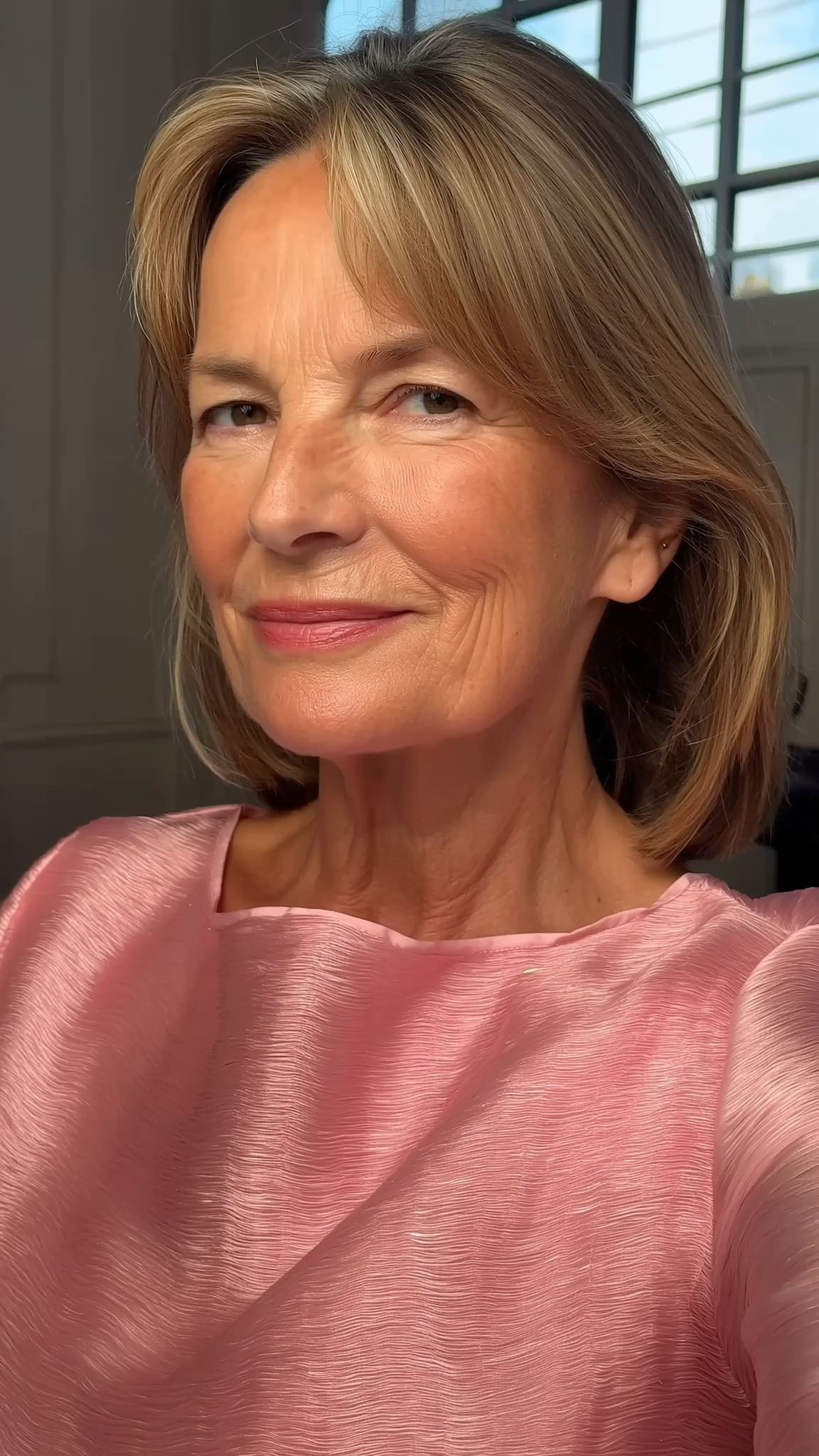 A smiling, middle-aged woman with blonde hair, wearing a shimmering pink top and looking at the camera.